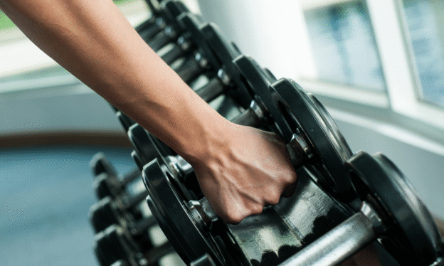 Close-up of a person’s hand gripping a dumbbell from a rack in a gym, with natural light from large windows.