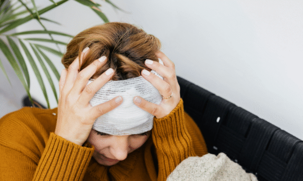 Woman with a head injury holding her head in pain, wearing a bandage and sitting on a couch in a mustard sweater.