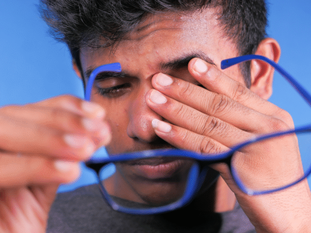 Close-up of a man removing his glasses and rubbing his eyes, showing signs of pain or eye strain.