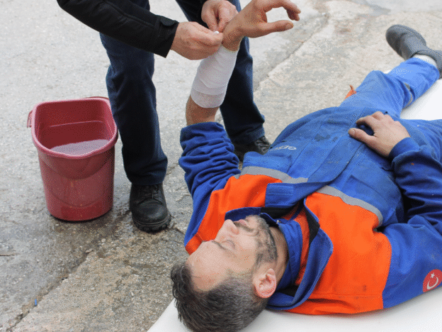 Injured worker lying on the ground in a blue and orange uniform while another person wraps his bandaged arm.